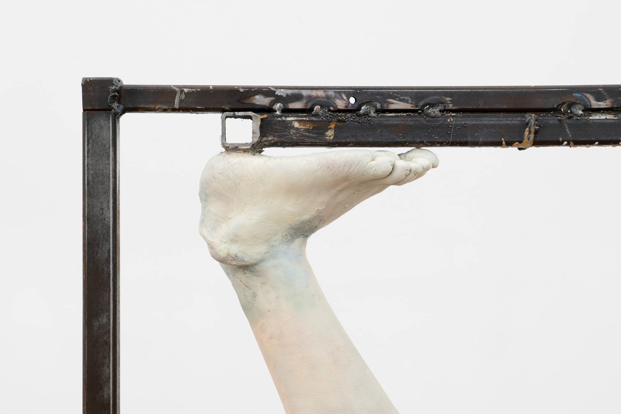 Detail of a freestanding sculpture featuring a fragmented figure, inverted and suspended from a metal frame.