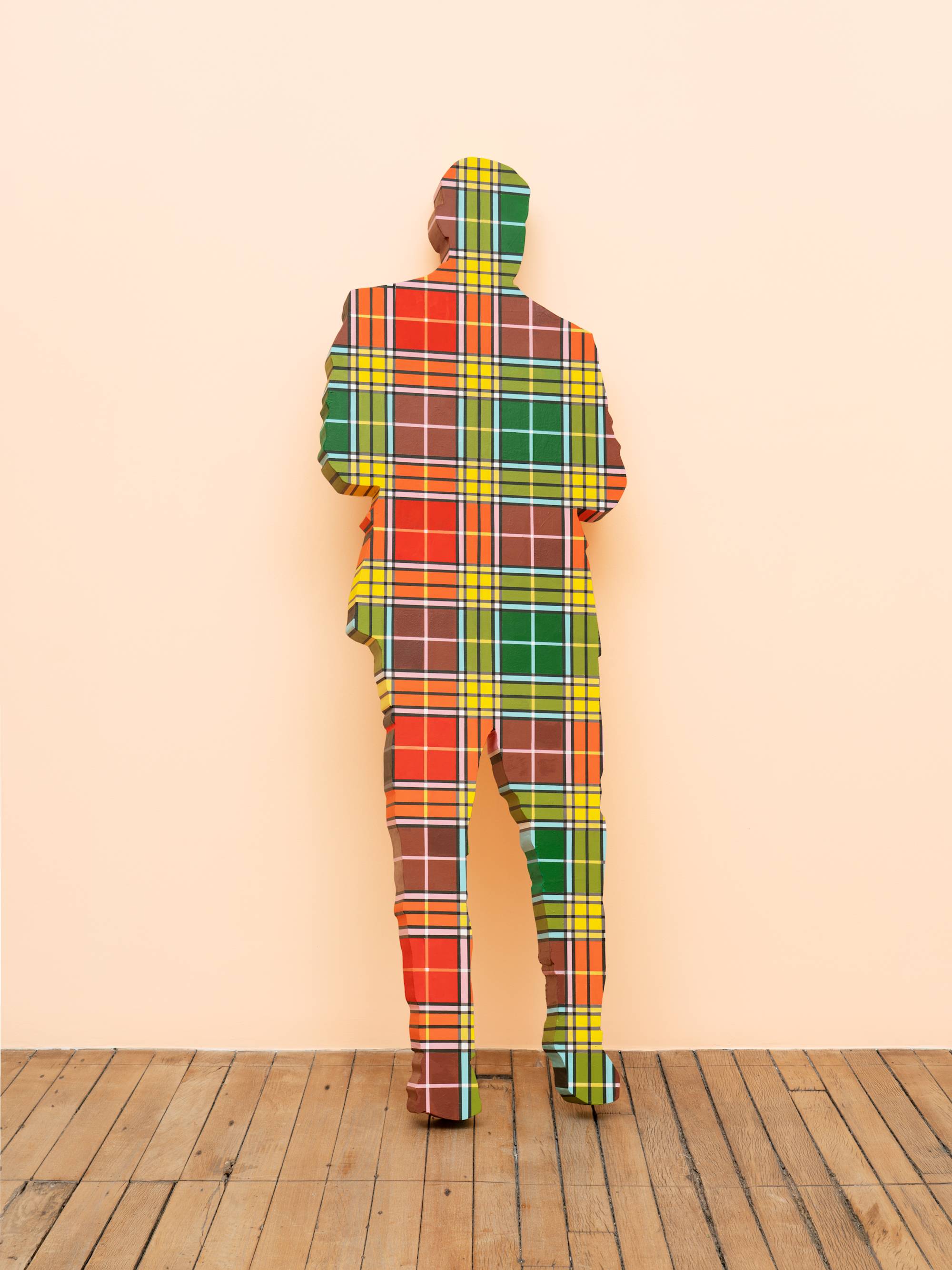 A life-sized human figure painted in a rainbow plaid pattern leans against a peach wall in an art gallery.