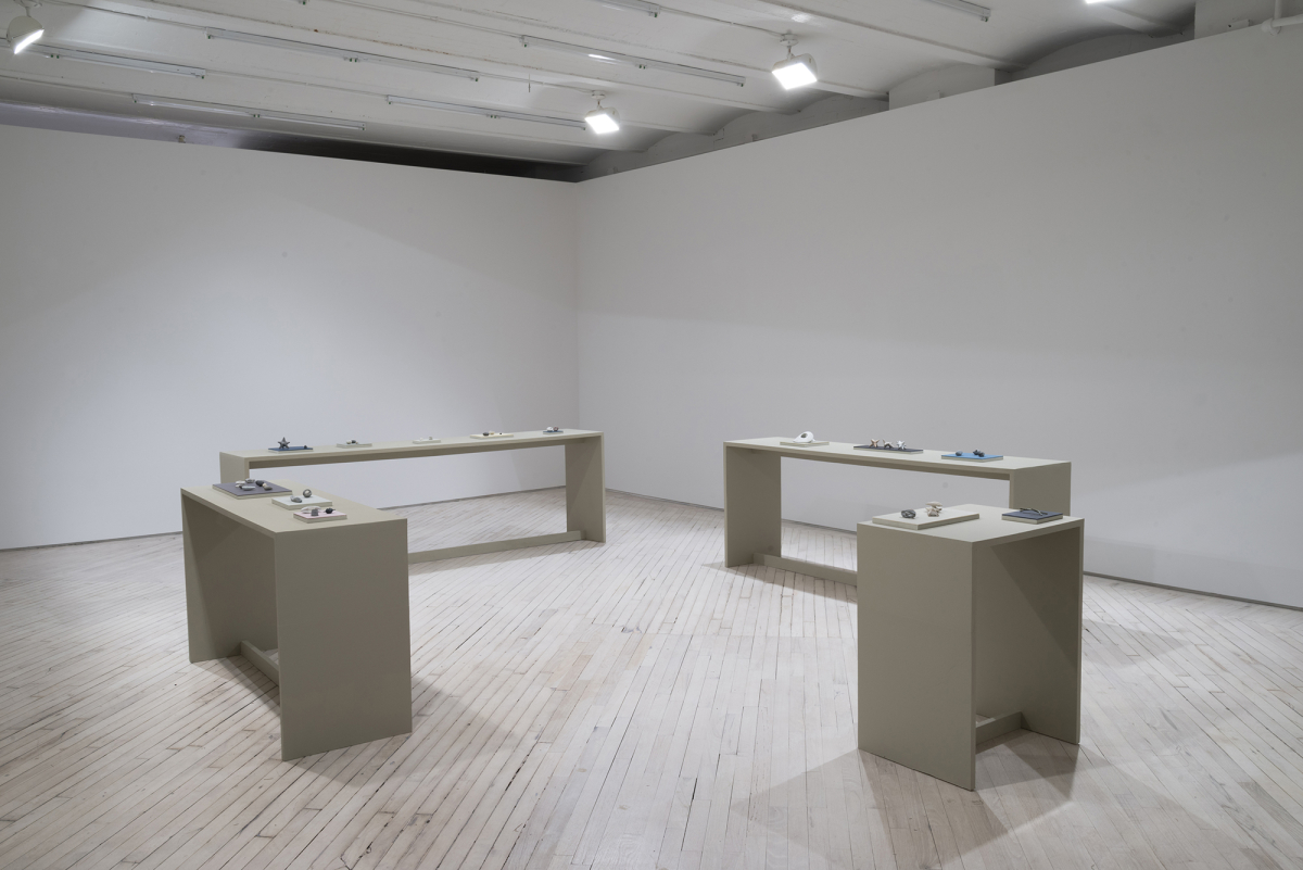 In a darkened gallery space, four tables arranged in a square facing one another. Resting on top of the tables are numerous small plinths housing various miniature sculptural objects. 