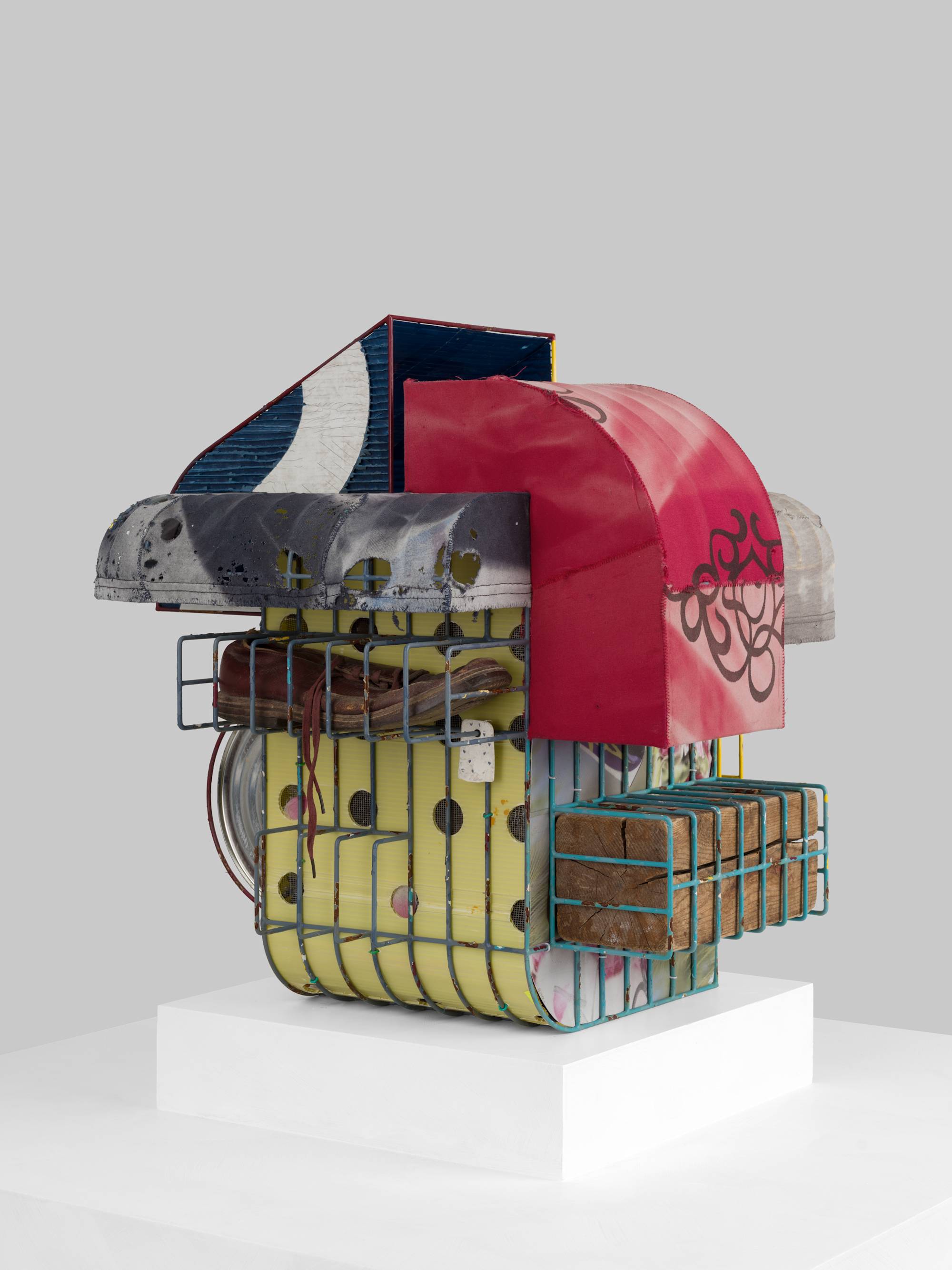 A cage-like colorful steal wire sculpture, wrapped with several tattered fabrics to resemble various awnings, and embellished with an old shoe, a twig, printed coroplast signage, an old blue paint can, a lock, a paper tag, and a wooden block sits on a white pedestal against a white wall.