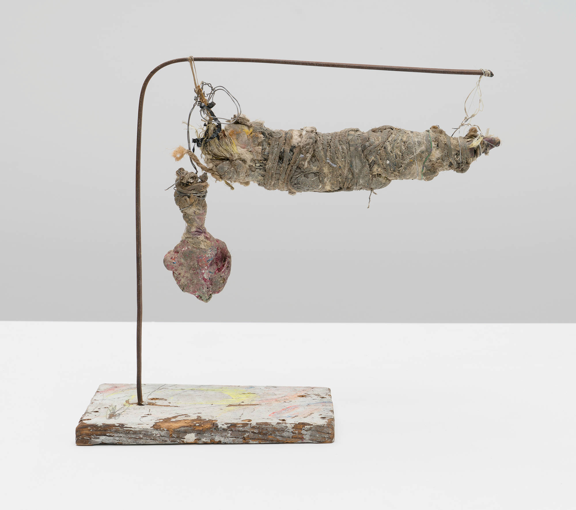 A small sculpture constructed of Tempera, ink, acrylic, crayon, graphite, string, cotton, wood, string, wire, plastic, metal, magnetic audio tape, beeswax, and mixed media resembles a mummy which is suspended from a metal arm attached to a wooden base.