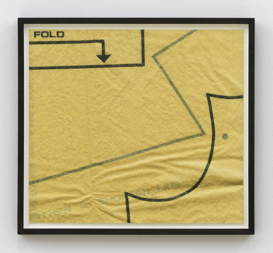 Image of a photograph in a black frame, the photograph depicts an up close look at a vintage sewing pattern. The pattern's paper is yellowed and wrinkled. The pattern is cropped and visible are a series of straight and curved black lines with the word "FOLD" at the top right corner above an arrow.
