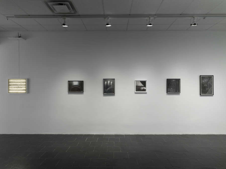 A series of charcoal drawings in black frames hanging in a line. On the left is a white lightbox containing a scroll of text.