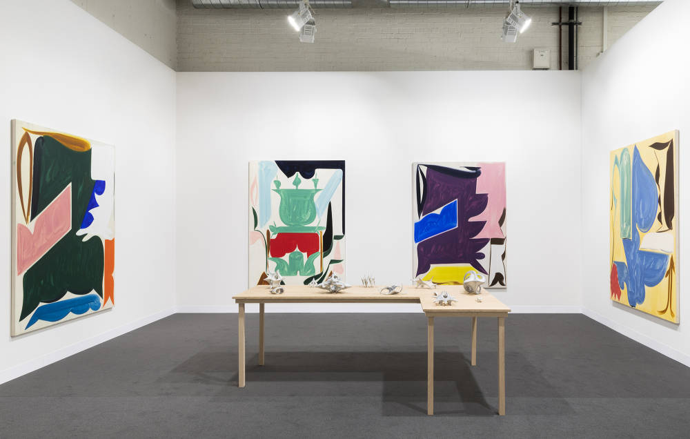 In a gallery space, four large abstract paintings are spaced evenly throughout. The painting depict bold abstract geometric shapes in various hues of color. The paint in the paintings is thin and gestural. In the center of the gallery space is a freestanding brown table with various miniature gray sculptures placed evenly apart. The sculptures are abstract and resemble organic material or bone formations. 