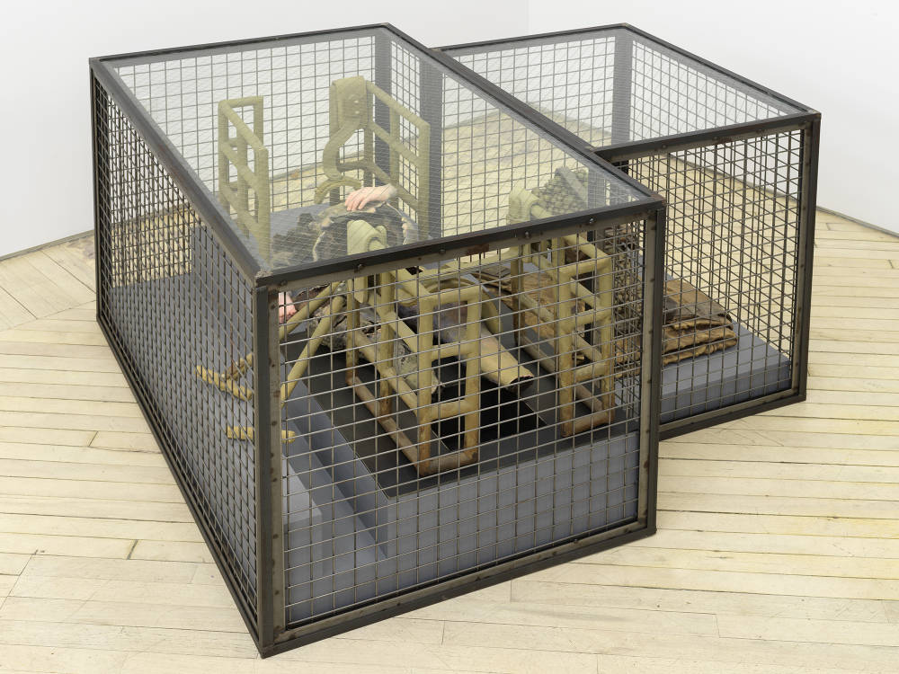 In a gallery space, a large iron sculpture resembling a cage containing another restriction device.