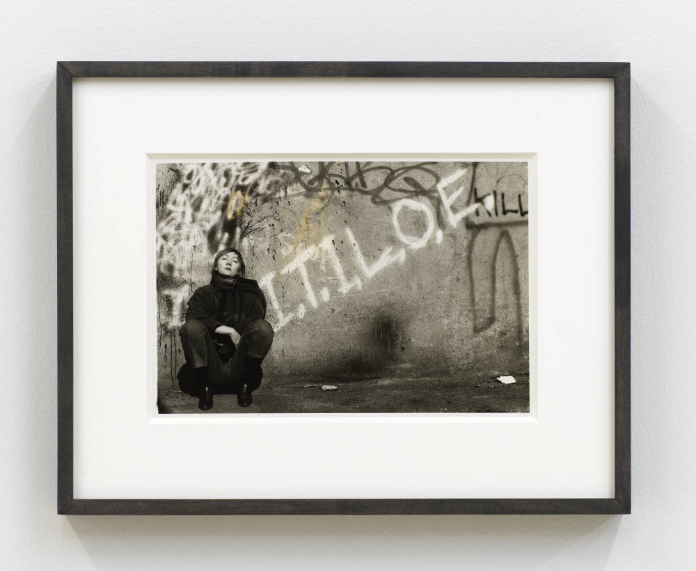 A small black and white photograph of a woman squatting with the text "I.T.I.L.O.E" painted on a wall. 