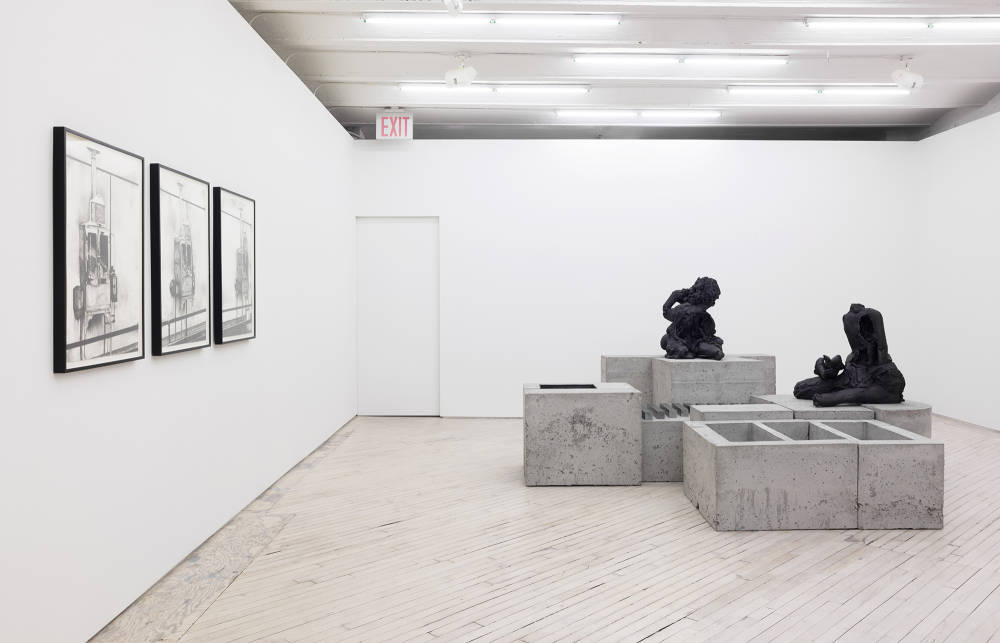 Installation view of a triptych of black framed black and white drawings on the left and a large grey and black floor sculpture at left and center.