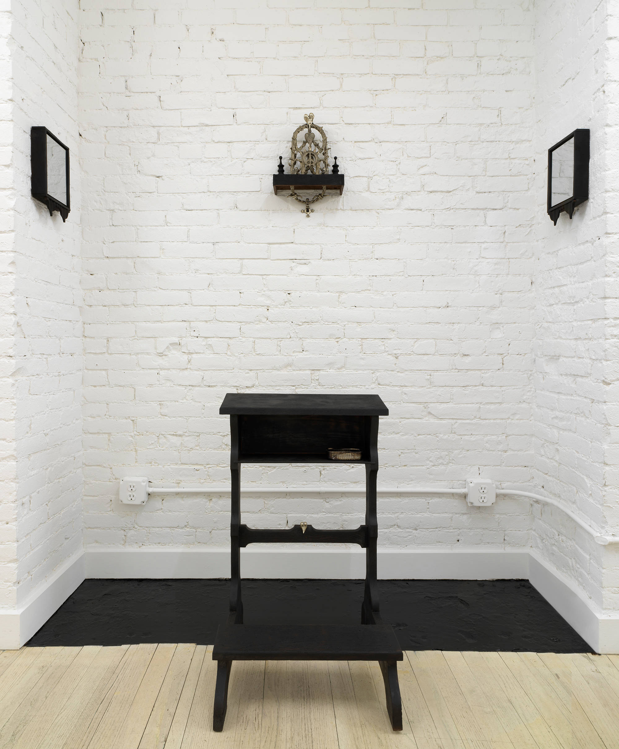 Installation view of Harvey's sculptural installation in a small niche in the hallway wall comprising a black chapel kneeler and three wall works hanging on the brick wall. The central work is an ornate cast bronze gothic style object featuring arcs and hearts and pointed elements. On left and right are 2 mirrors framed in black wood, which face one another.