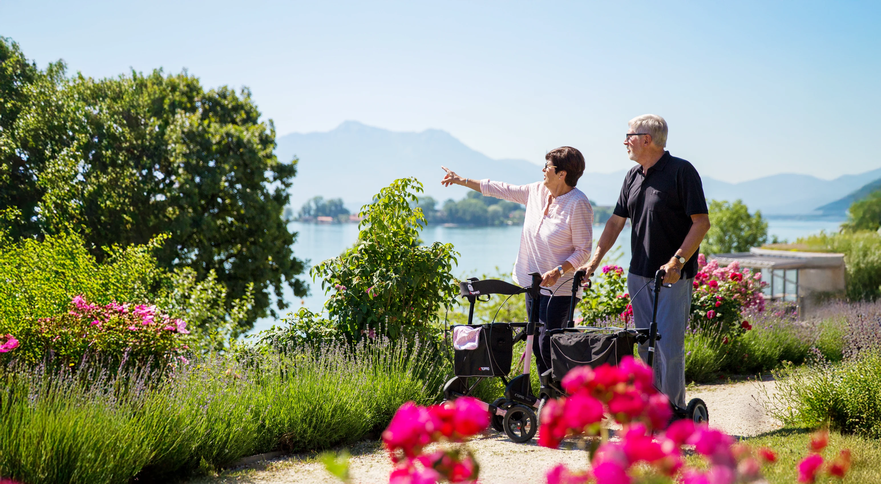 Two people using walkers in a garden area, one pointing toward a lake and mountains in the distance.
