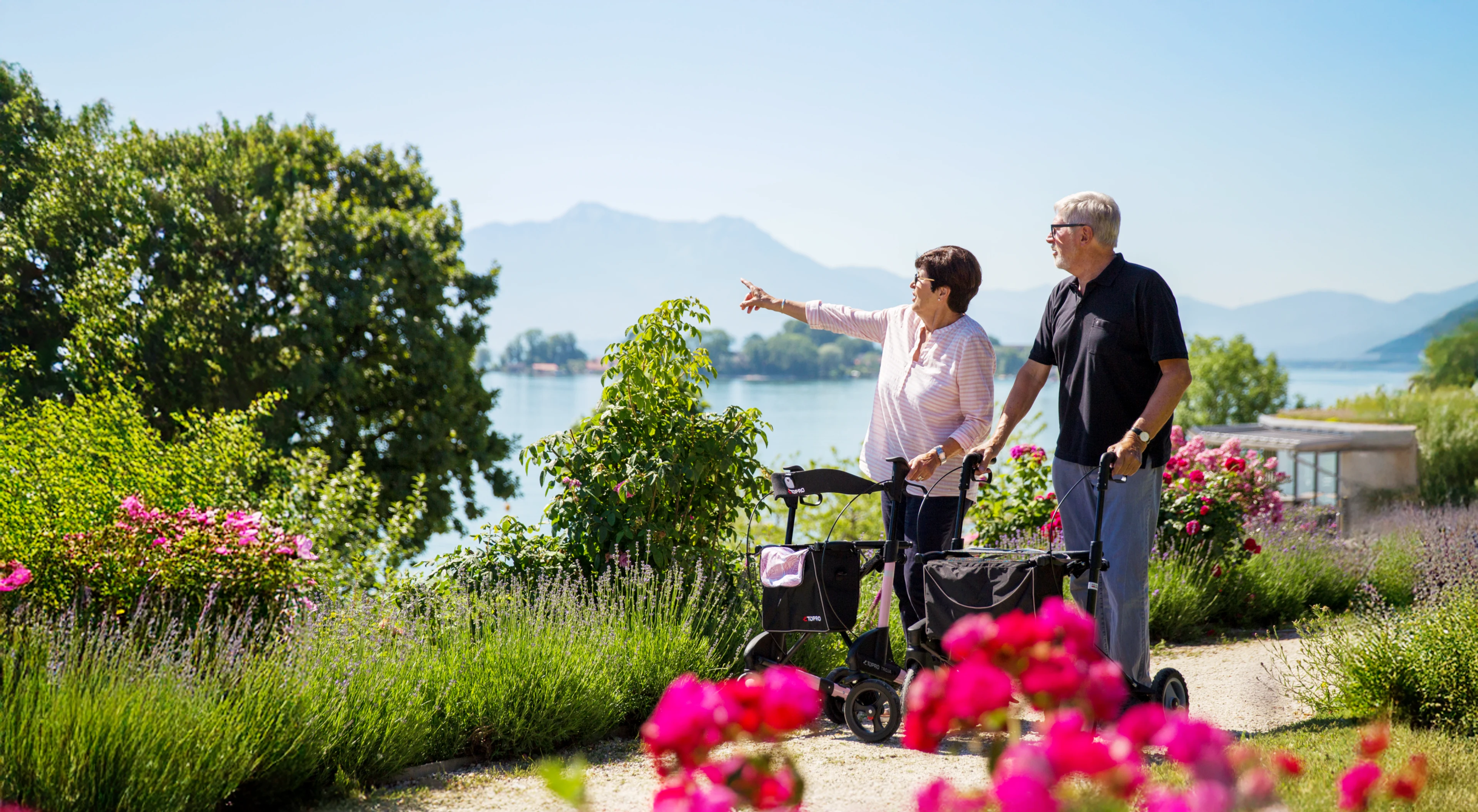 Two people using walkers in a garden area, one pointing toward a lake and mountains in the distance.