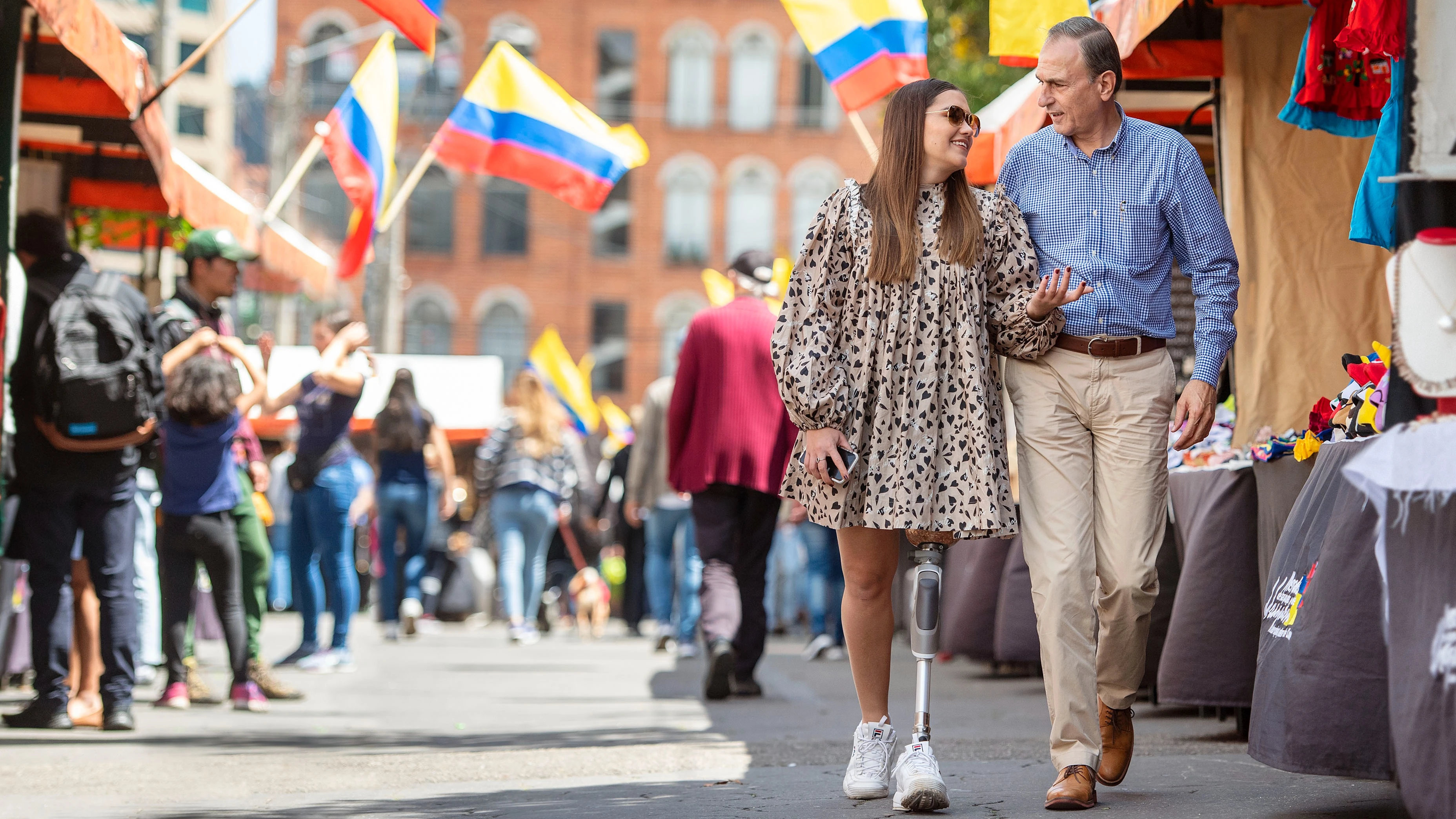 Una pareja caminando en terreno plano a través de un mercado al aire libre. Uno de ellos lleva la rodilla mecánica Ottobock Dynion
