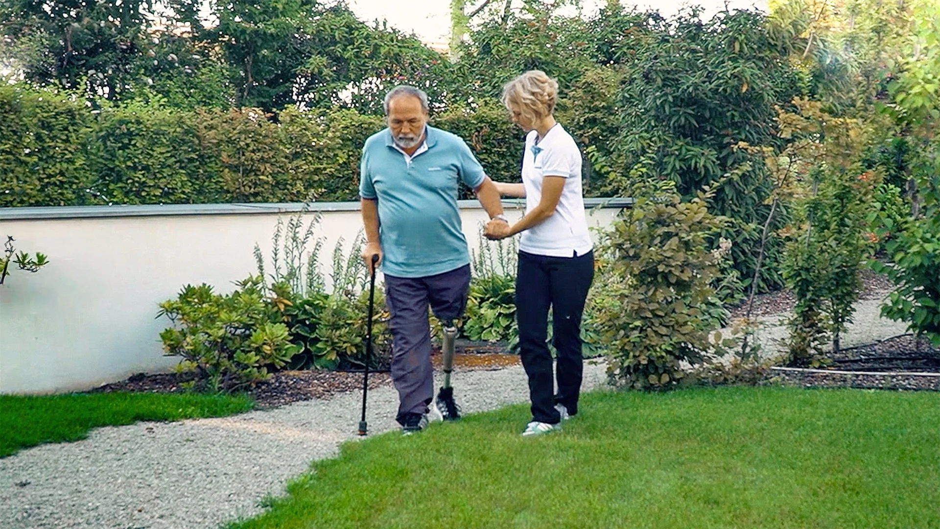 An Ottobock Kenevo knee prosthetic user walking outdoors on an asphalt walkway with assistance