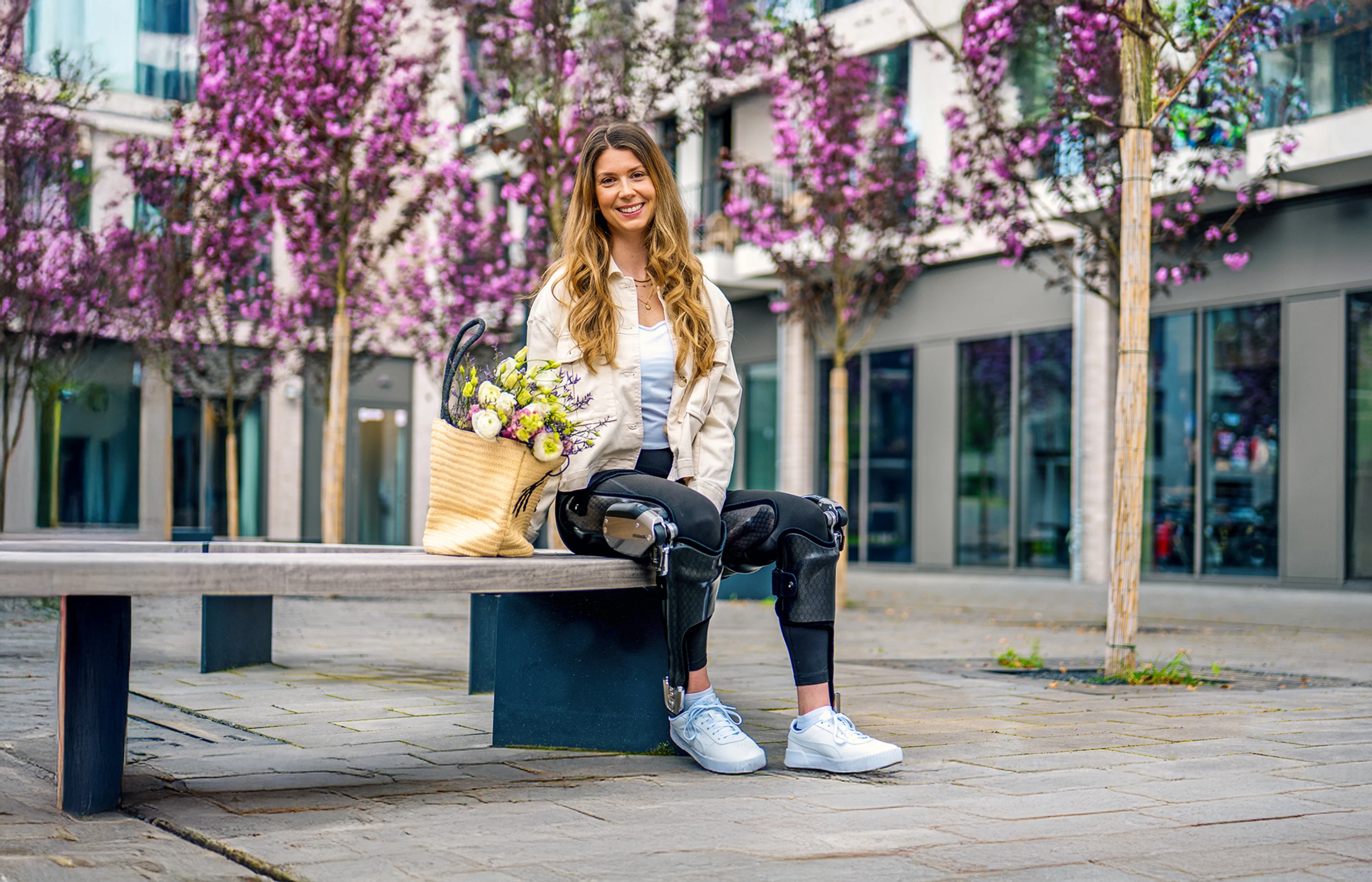 Person sitting on a bench in an urban area wearing leg orthotics on both legs, with flowers beside them.
