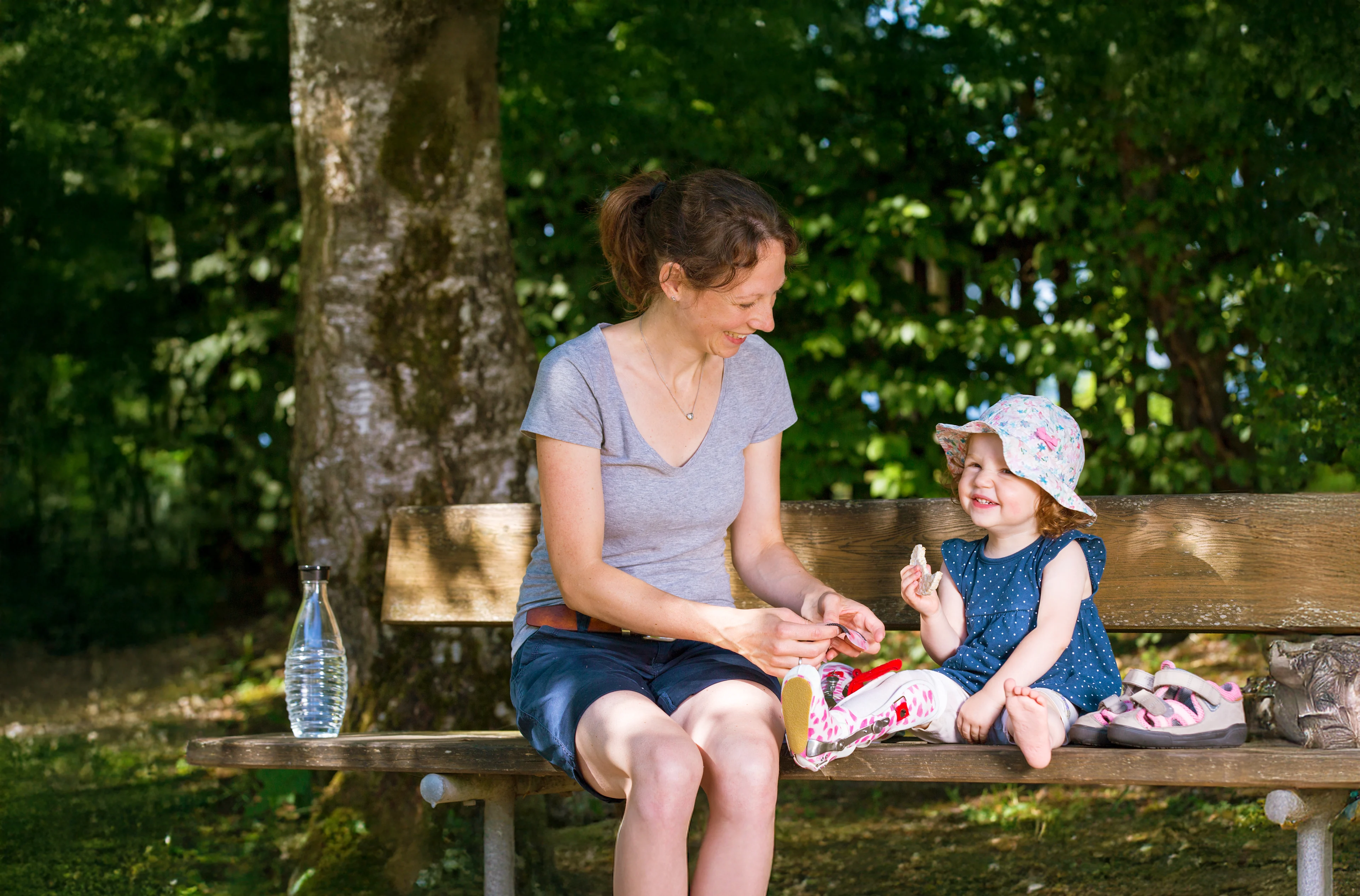 Adult and child sitting on a wooden bench in a park; the adult is handing something to the child.