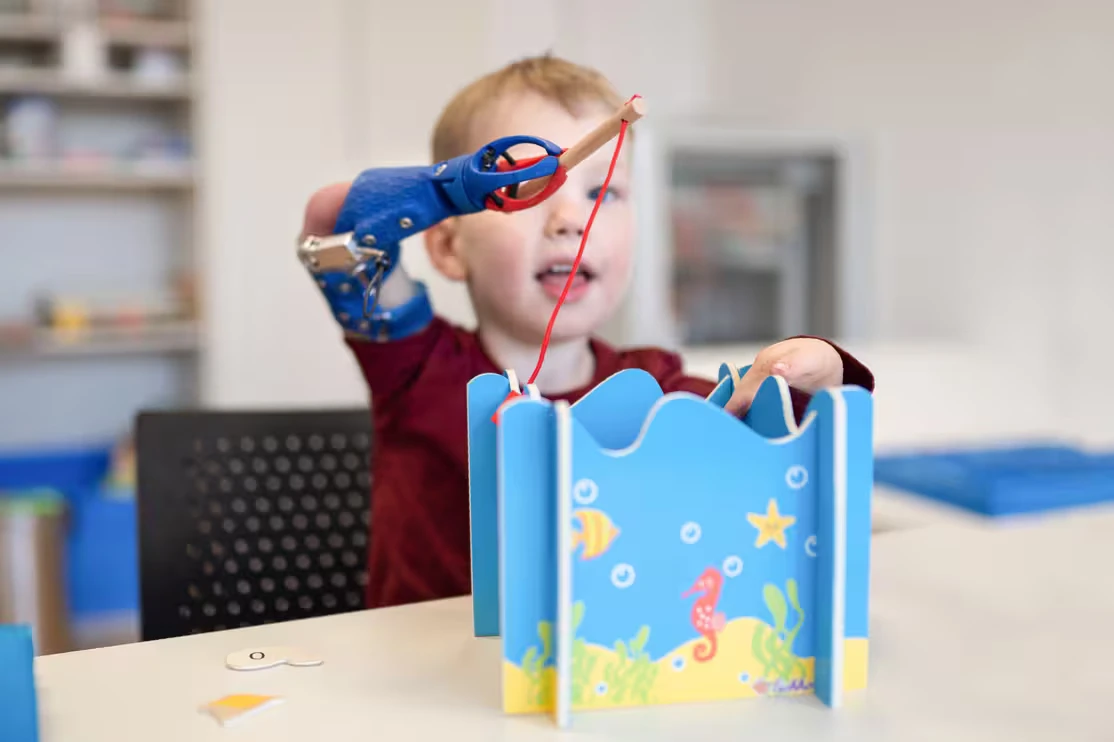 Child using a prosthetic arm to play a toy fishing game indoors.