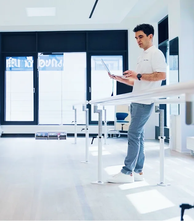 Person standing at parallel bars using a laptop in a rehabilitation setting.