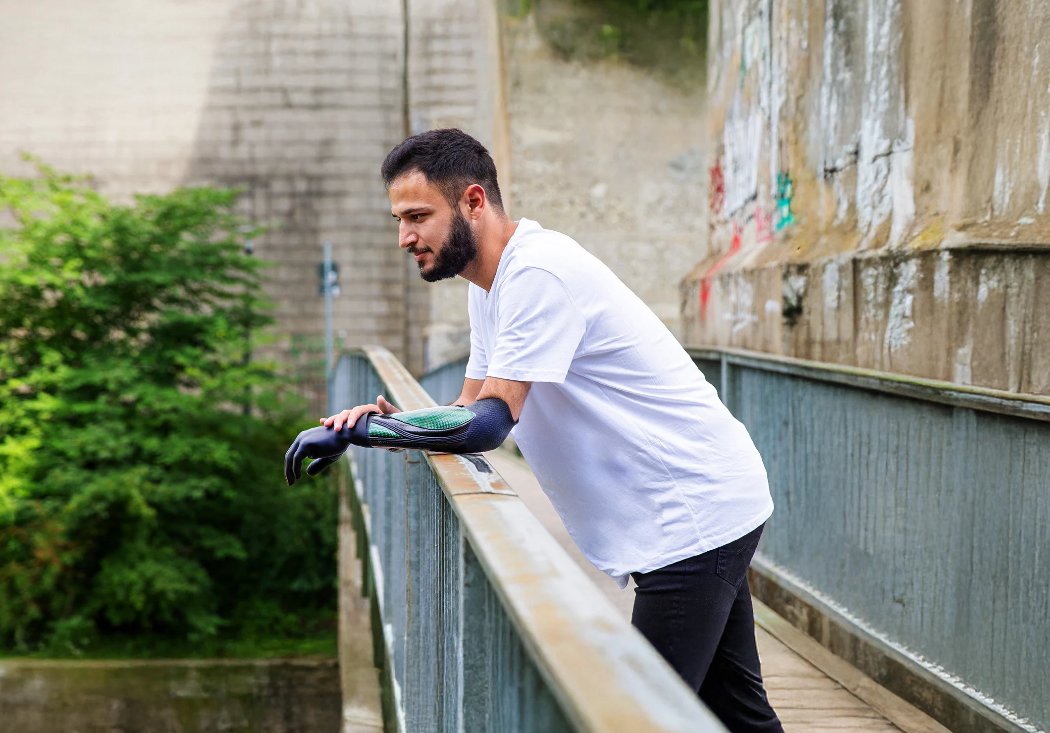 Person leaning on a bridge railing, looking downward, with greenery in the background.