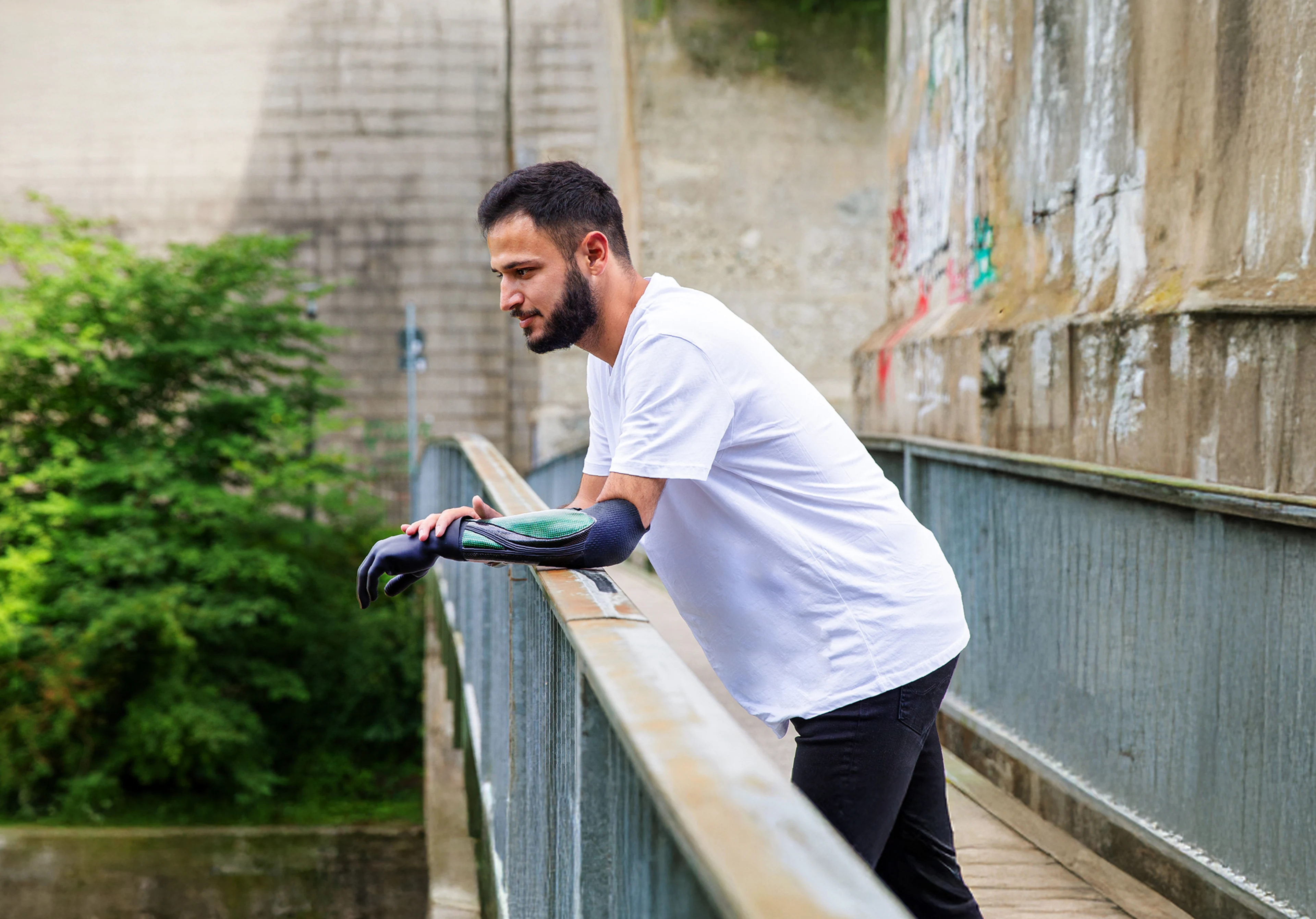 Person leaning on a bridge railing, looking downward, with greenery in the background.