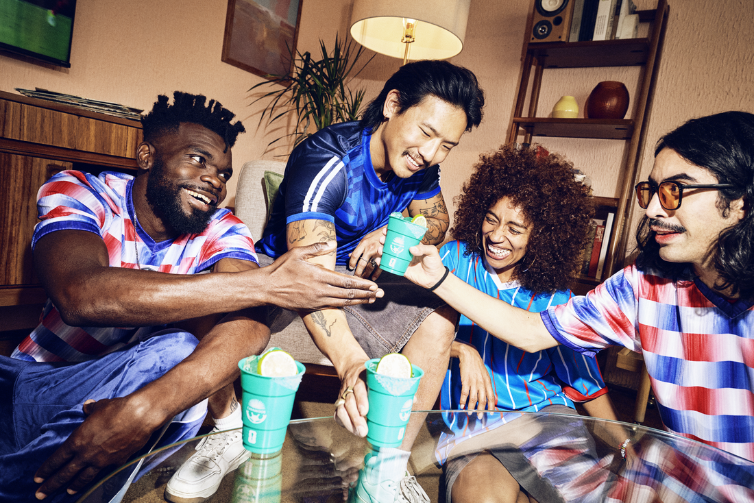 Friends wearing football jerseys gathered around a table at home, smiling and laughing while enjoying margaritas during a World Cup watch party.