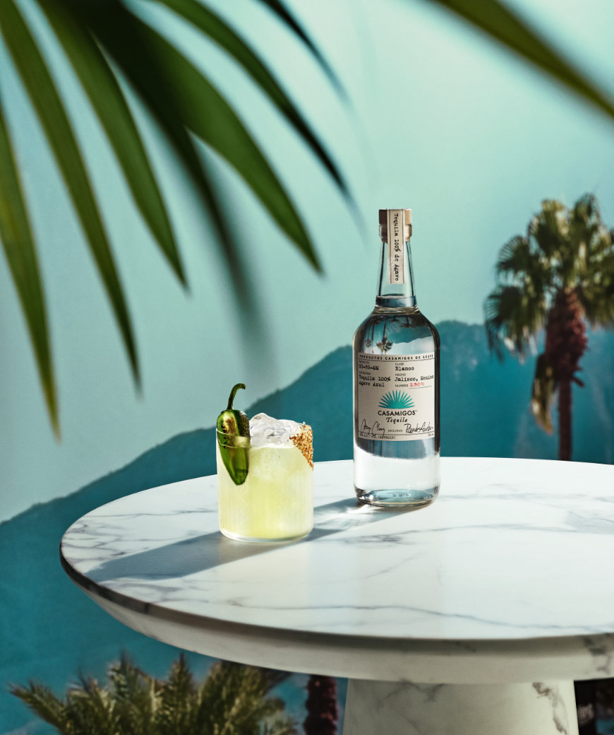 A Casamigos Spicy Margarita cocktail with half slice of jalapeno garnish next to a bottle of Casamigos Blanco tequila.