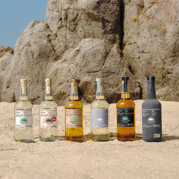 Collection of Casamigos tequila bottles: Blanco, Jalapeño, Reposado, Cristalino, Añejo, and Mezcal, arranged on sand with a rocky coastal backdrop.