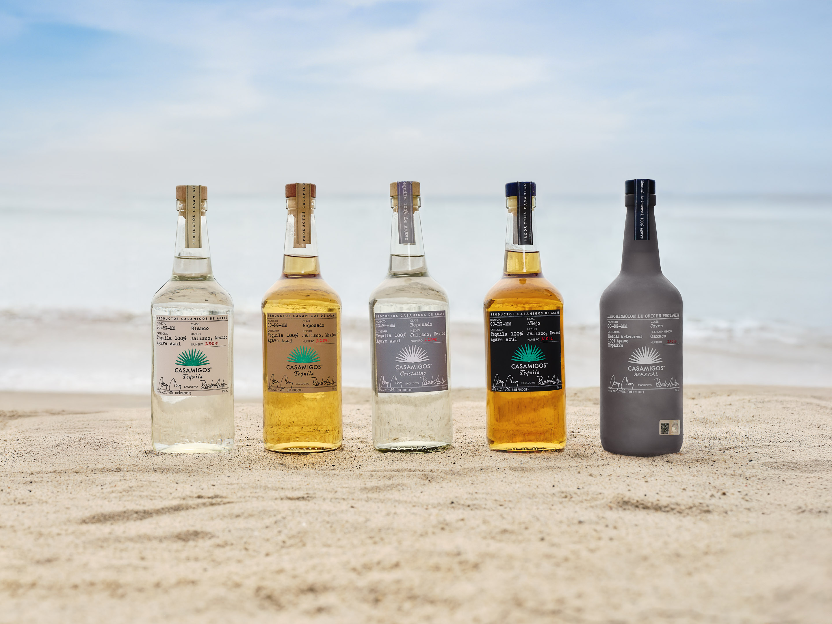 Collection of Casamigos tequila bottles: Blanco, Jalapeño, Reposado, Cristalino, Añejo, and Mezcal, arranged on sand with a rocky coastal backdrop.
