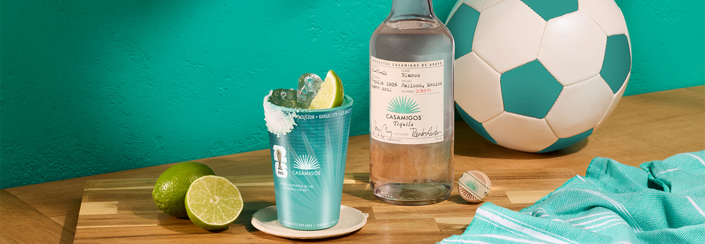 Bottle of Casamigos Blanco with a margarita on the rocks in a commemorative cup featuring a salted rim, set on a butcher block beside a teal soccer ball.