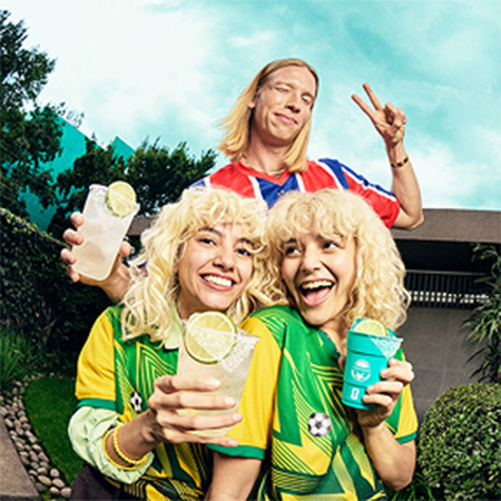 Twins in yellow-green football jerseys with margaritas, friend in red-blue jersey, celebrating at a World Cup watch party.