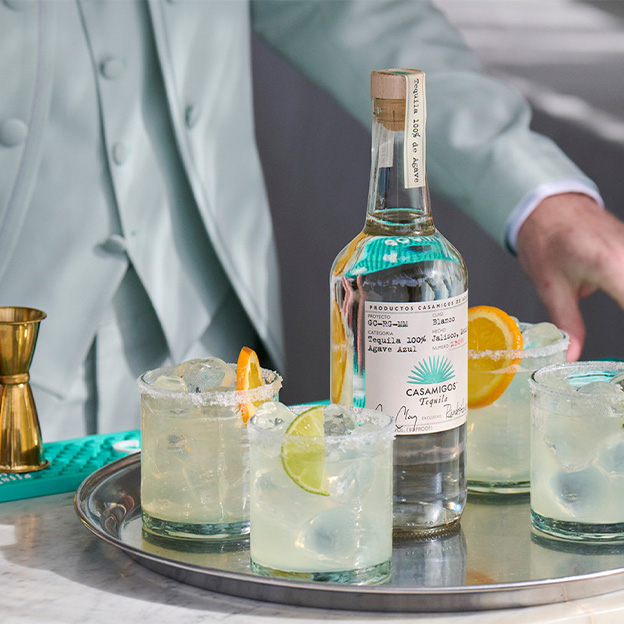 Casamigos Blanco tequila bottle surrounded by classic margarita cocktails on the rocks with fresh lime and orange garnish on a modern bar tray