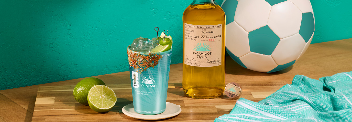Bottle of Casamigos Blanco with a margarita on the rocks in a commemorative cup featuring a spicy salted rim, set on a butcher block beside a teal soccer ball.