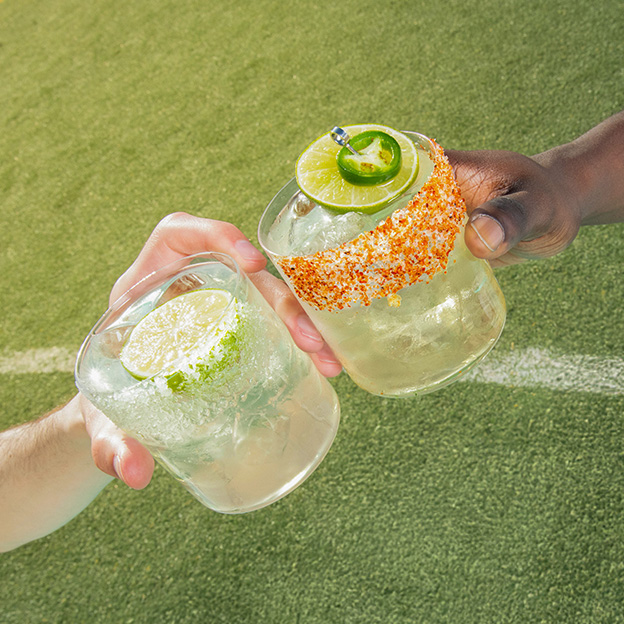 Two hands clink margaritas on the rocks over a soccer field line — one classic with a salted rim, the other spicy with a chili-salt rim — set against green turf.