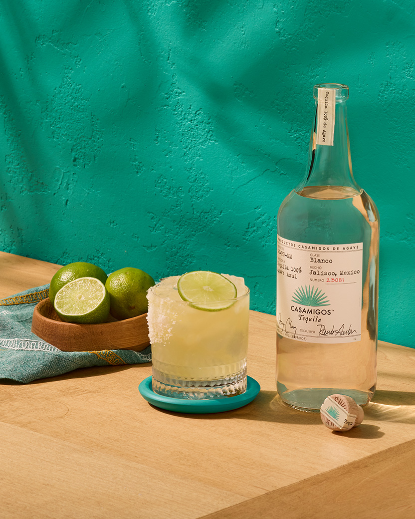 A Casamigos Margarita in a rocks glass with lime garnish on a small platter with a bottle of Casamigos Blanco tequila.