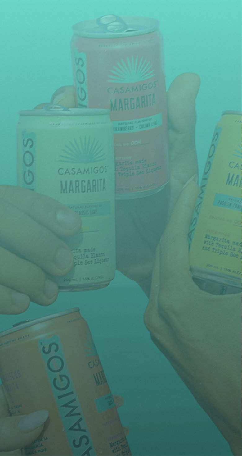 Four friends raising Ready-To-Drink Casamigos Margaritas cans in a toast with a teal ocean and sky in the background