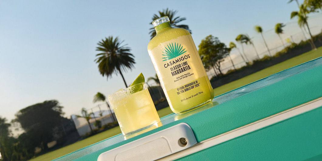 Casamigos Classic Margarita bottle and poured margarita in a rocks glass on a teal cooler, set against tropical palm trees.
