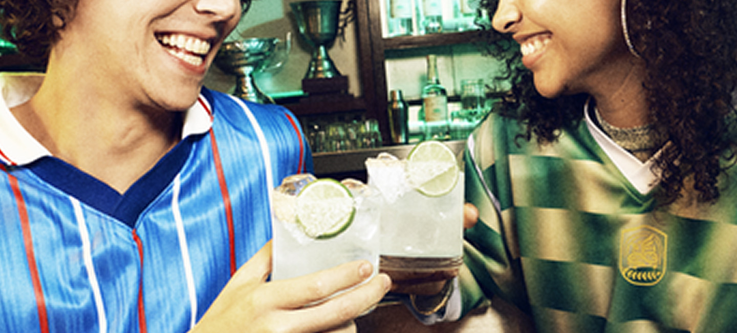 Two friends in different football jerseys smile and clink margaritas on the rocks at a bar, with two trophies and tequila bottles visible in the background.