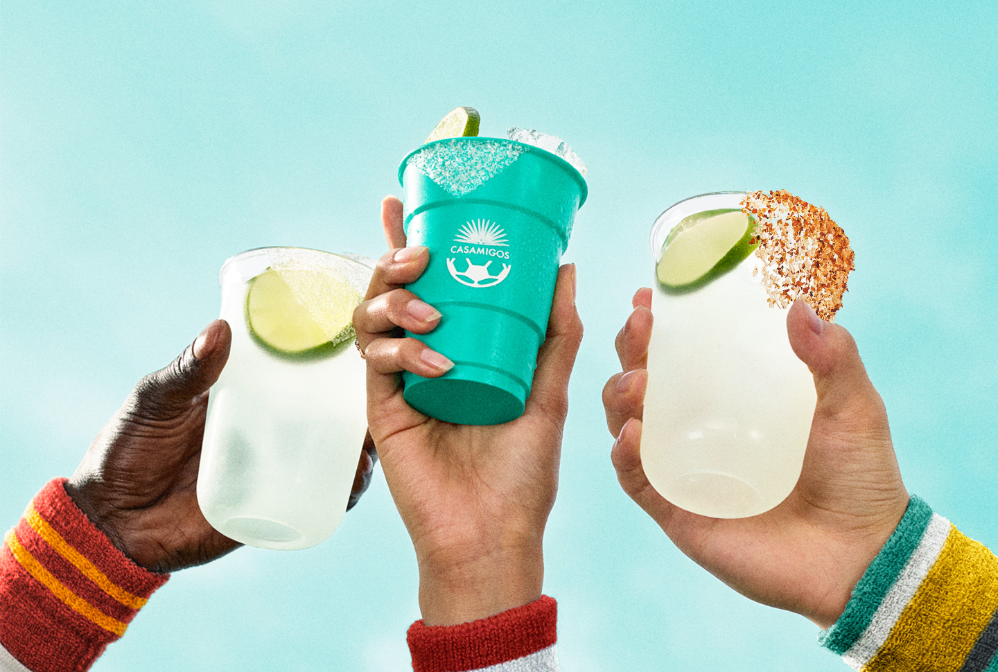 Friends toasting margaritas with lime wedges in teal and clear drink cups beneath a bright blue sky.