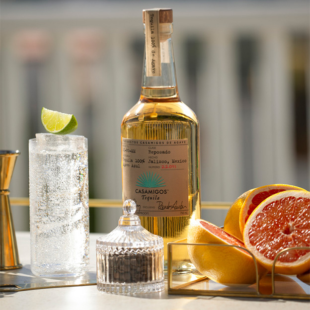 Casamigos Reposado tequila bottle on a tray with a jigger, clear sparkling cocktail in a Collins glass, and fresh grapefruit halves
