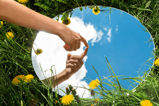 nature concept - hand touching sky reflection in round mirror on summer field