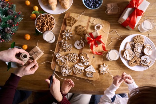 Top view from above of family and kid daughter making family xmas tree holiday gingerbread decorations holding Christmas cookies in hands preparing gifts together on festive decorated table, close up.