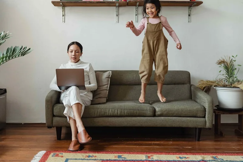 mom sitting on couch while child jumps on the couch