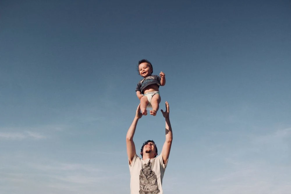 A man lifts a smiling baby high overhead against a clear blue sky.