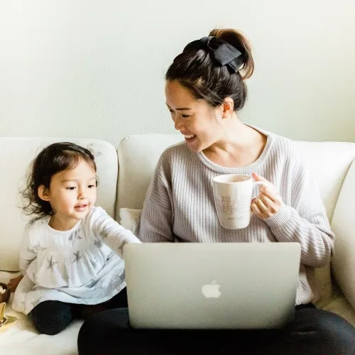 Mother drinking coffee and looking at custodianship while her baby is playing on the couch.