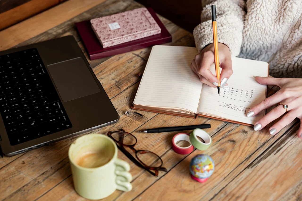 A person writing in a notebook at a wooden desk, with a laptop, coffee mug, and glasses nearby.
