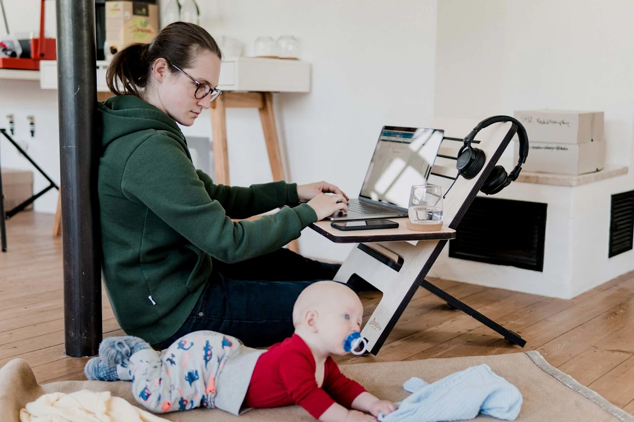 A parent sits on the floor working on a laptop at a low desk while a baby lies on a blanket nearby.