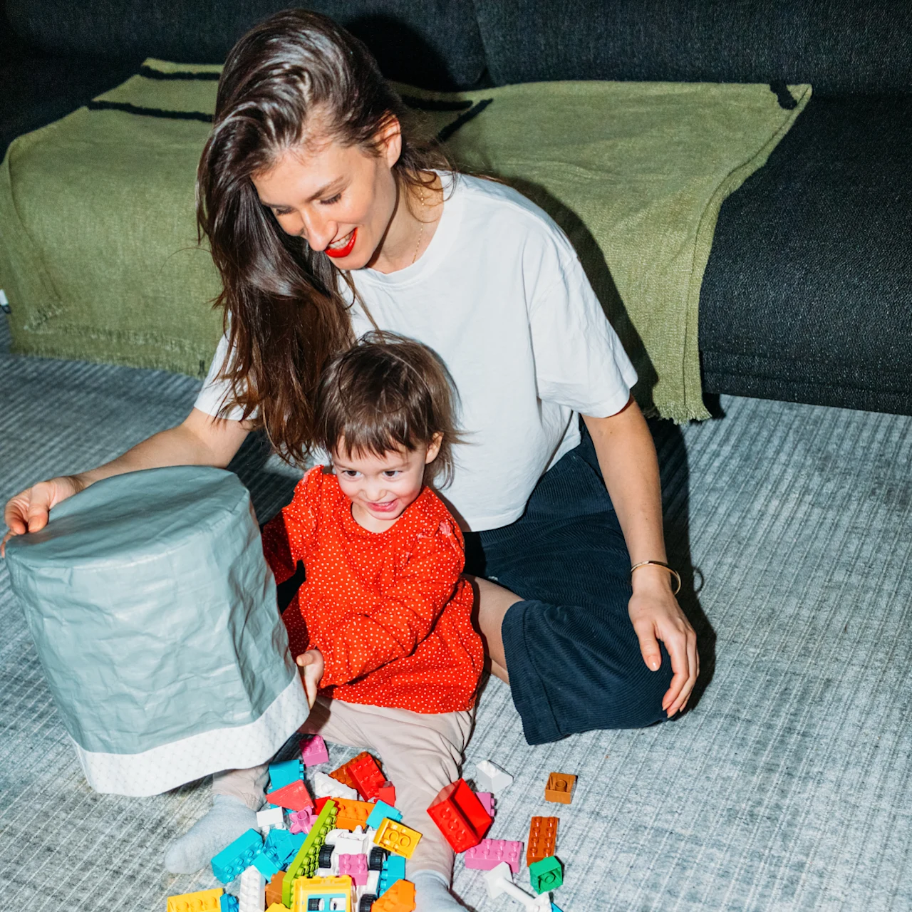 A smiling mother sits on the floor with her toddler, surrounded by colorful building blocks.