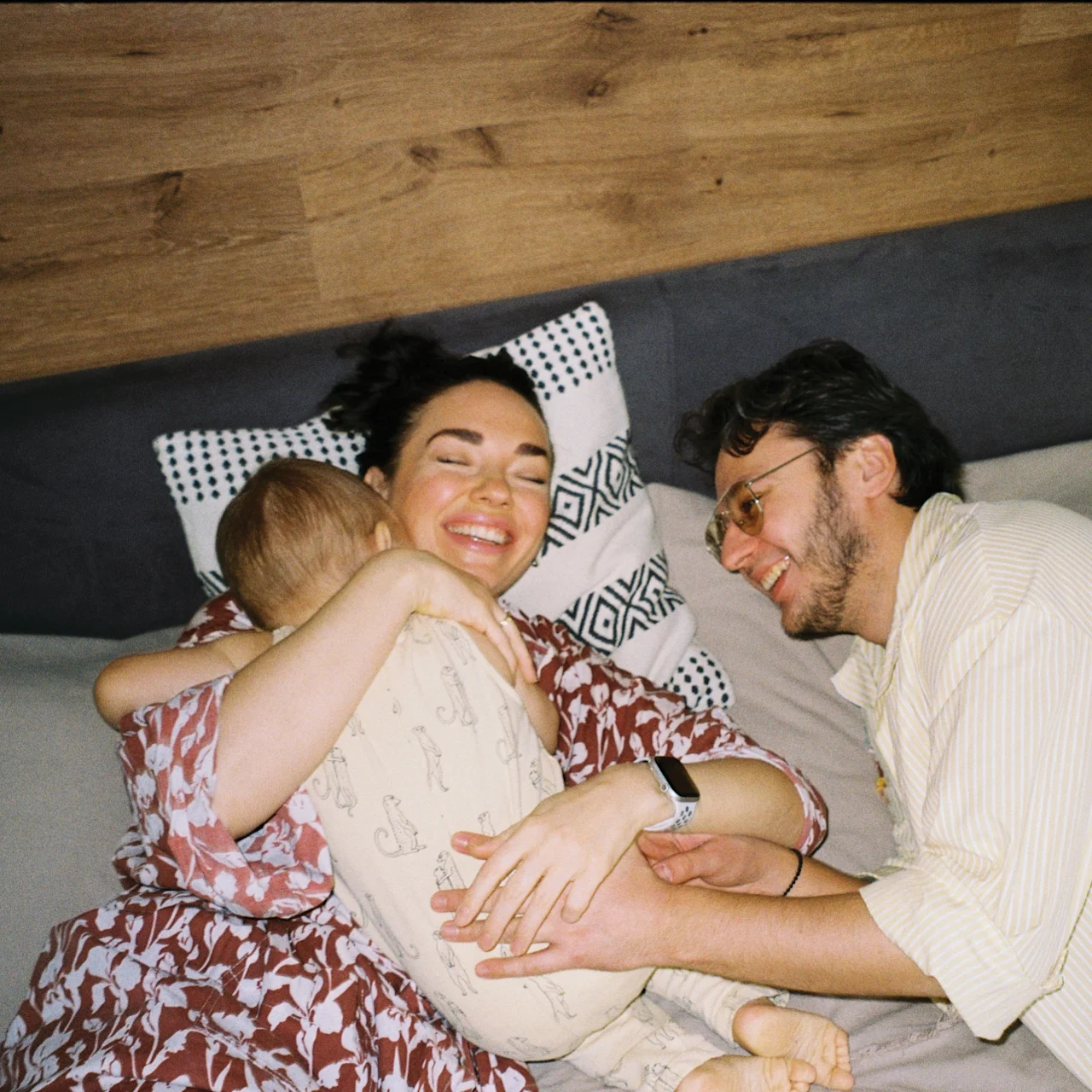 A mother, father, and baby laugh together on a bed.