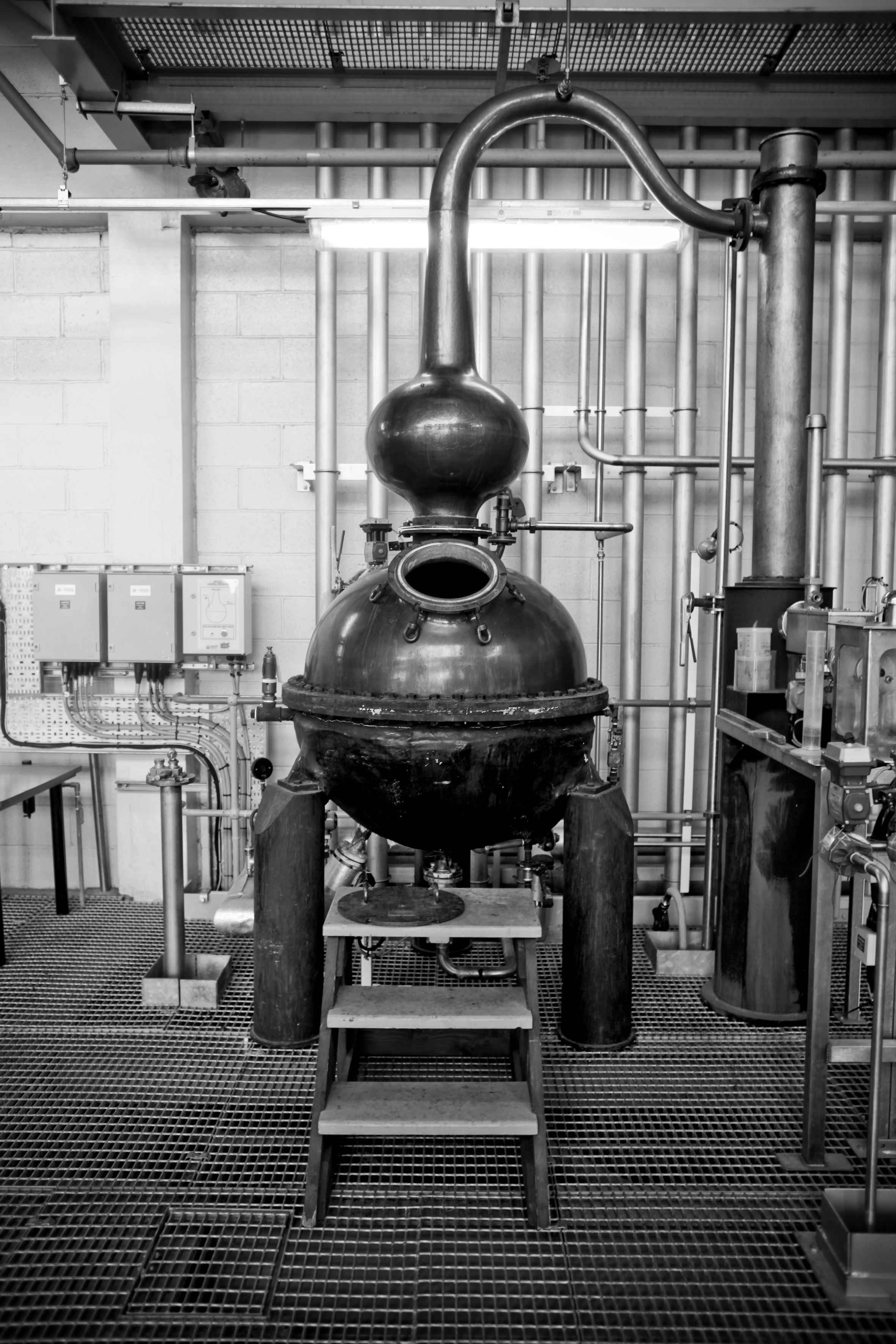 Black-and-white photo of a large metal distillation still with pipes and control panels in an industrial workshop.