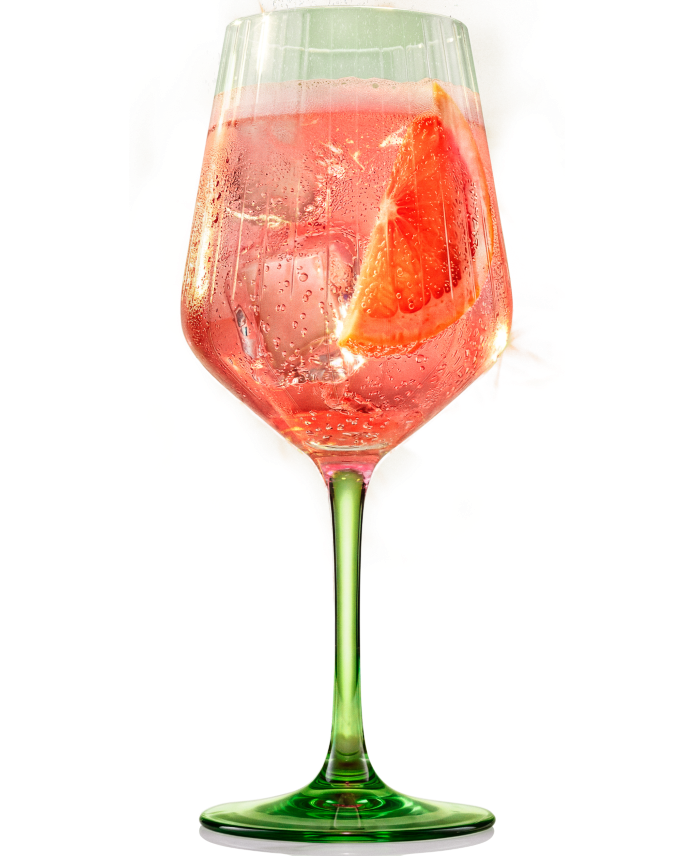 A ribbed, stemmed wine glass filled with a sparkling pink cocktail, ice cubes, and a slice of fresh blood orange or grapefruit. The glass features a bright green stem and base against a dark background with golden light reflections.