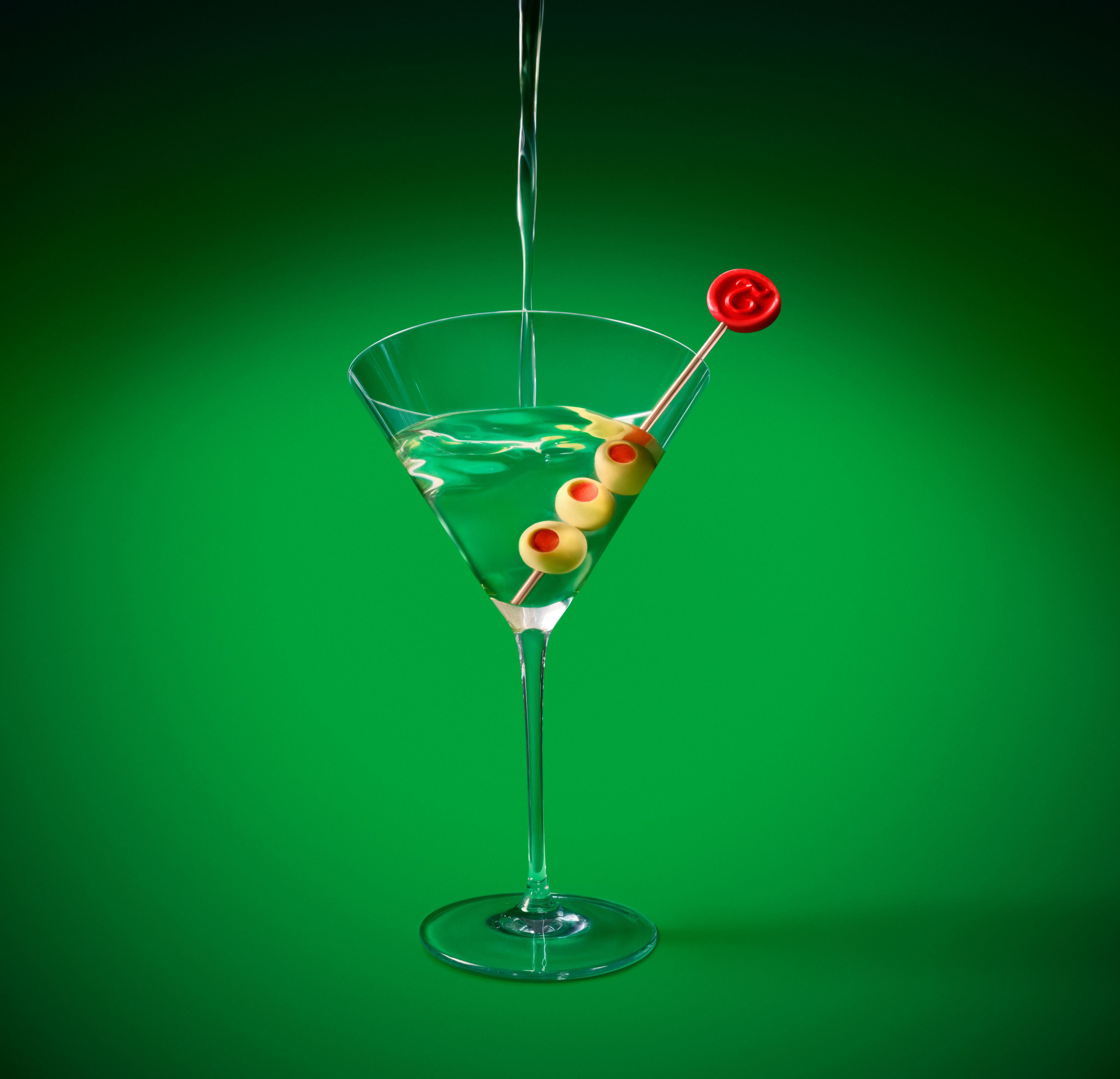 Clear martini glass with liquid being poured, garnished with three olives, on vivid green background.