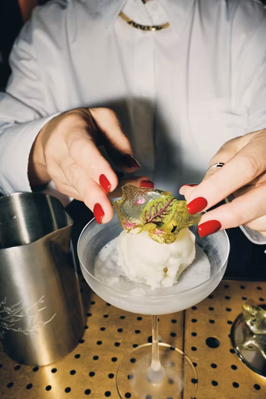 Person with red nails placing decorative sugar garnish onto sorbet in a coupe glass beside metal pitcher on perforated table.
